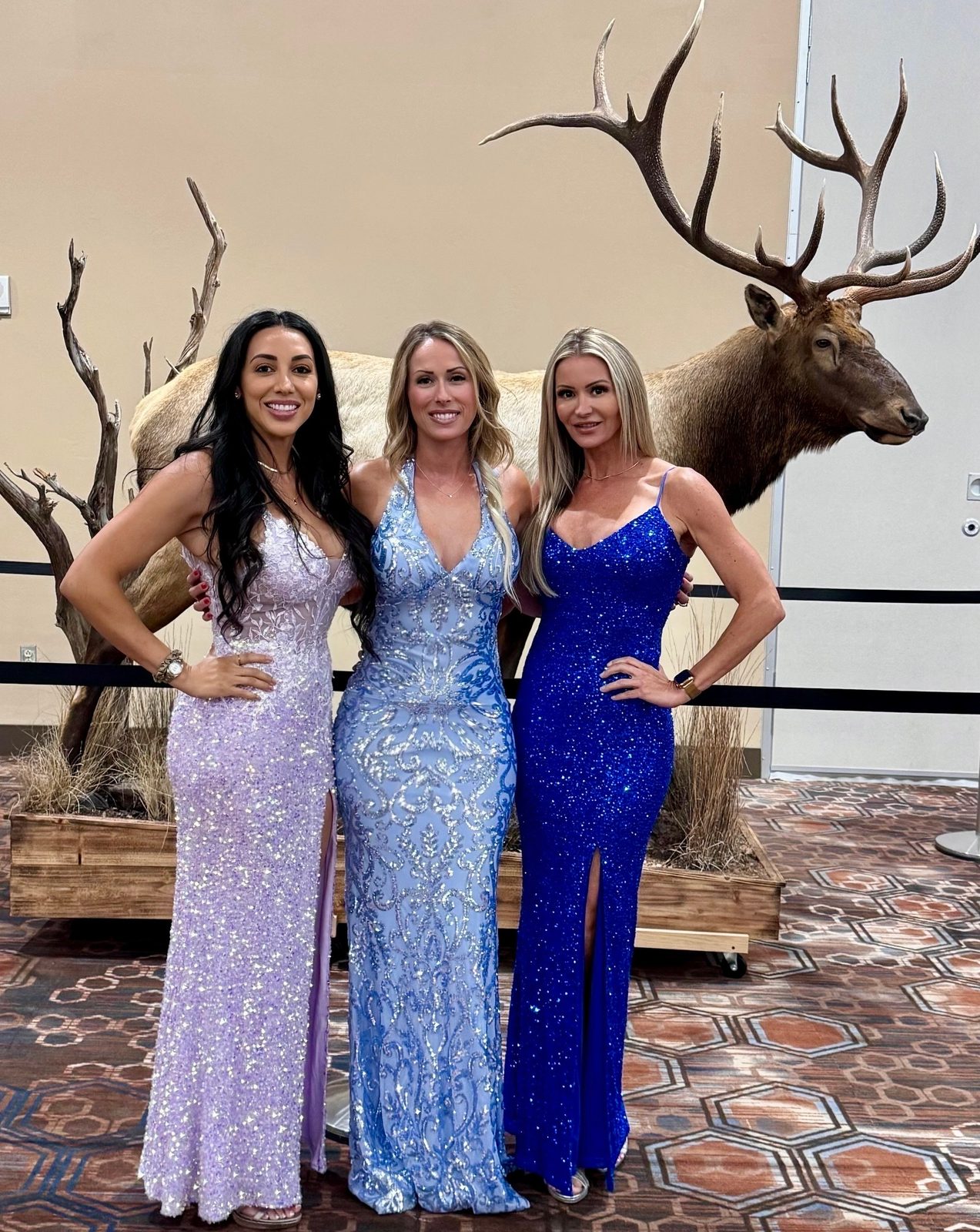 Three Mandi's Models in sequin gowns in front of a full-body elk mount