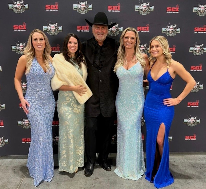 Four Mandi's Models in sequin and satin gowns with a guest in a cowboy hat at an SCI backdrop