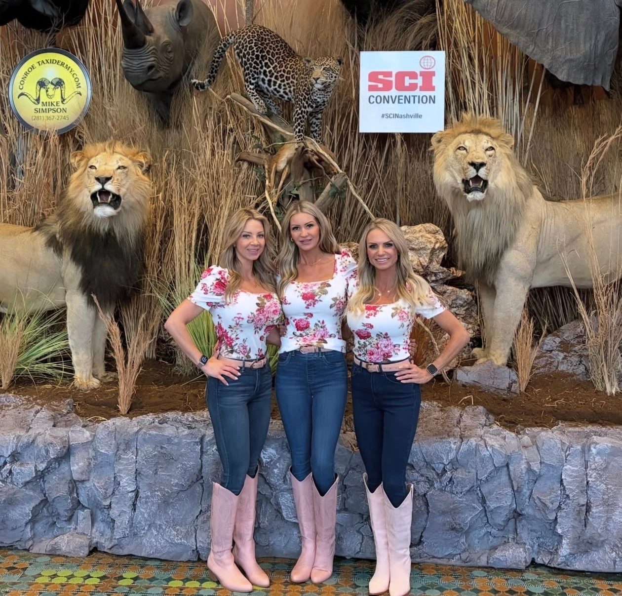 Three Mandi's Models in floral tops and pink cowboy boots at a lion display by an SCI Convention sign